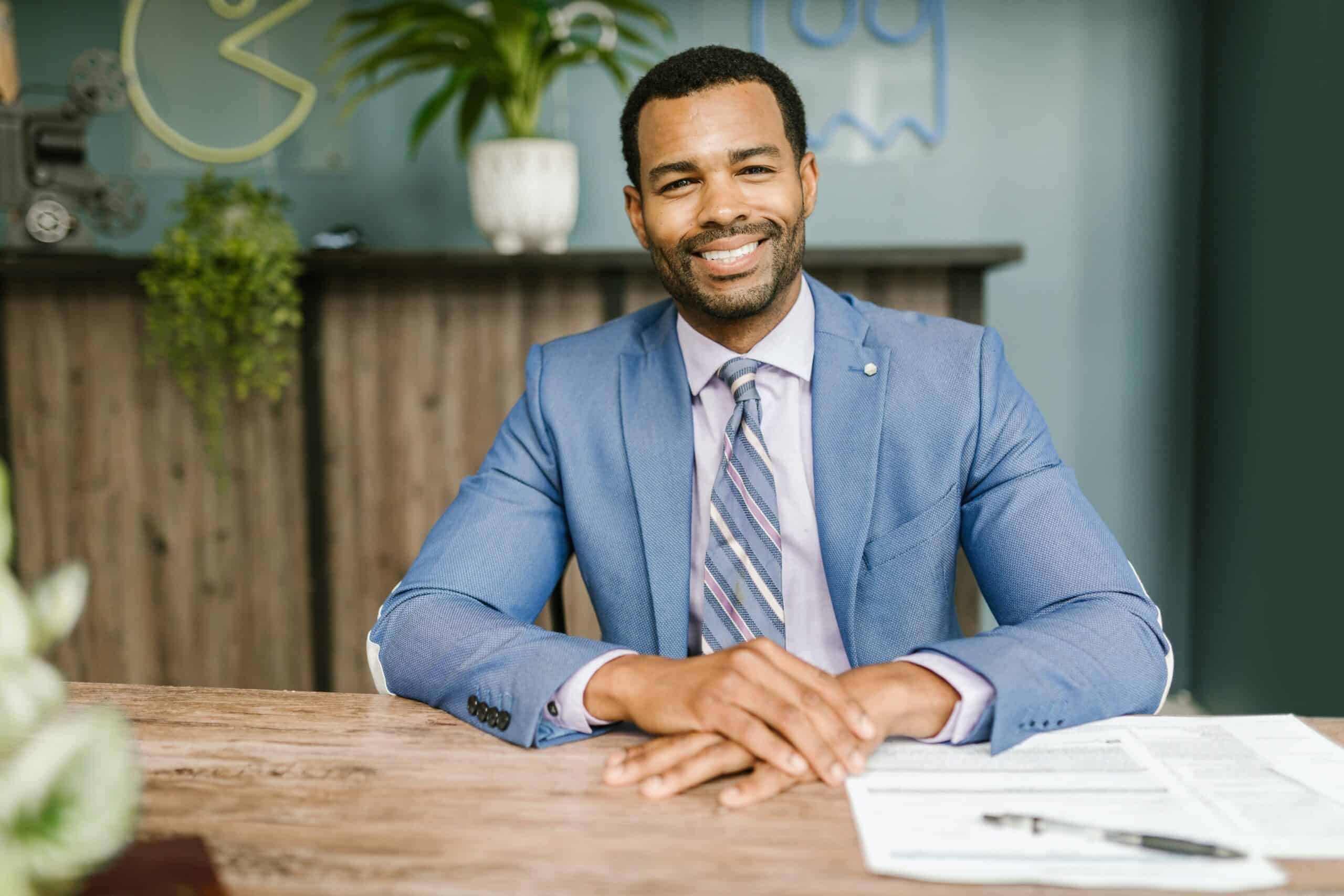 A smiling sharp dressed dark skinned man in sky blue suit sitting on a busy desk representing a forex broker